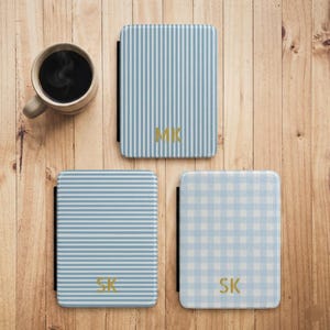 Puede incluir: Tres fundas de teléfono personalizadas con patrones en azul claro y blanco, incluyendo rayas verticales, rayas horizontales y un diseño de cuadros. Cada funda presenta iniciales doradas: MK y SK. Una taza de café está cerca sobre una superficie de madera.