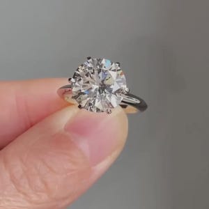 May include: A solitaire ring featuring a large, round, brilliant-cut clear gemstone held in a six-prong setting. The band is crafted from a smooth, polished silver-toned metal, providing a clean and reflective surface that complements the stone.