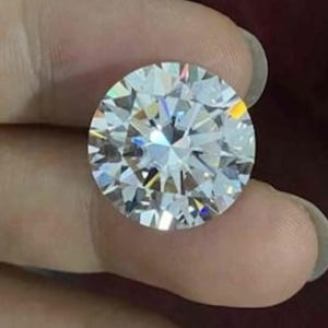 May include: A round, brilliant-cut, colorless gemstone featuring numerous triangular and kite-shaped facets that create a high level of light reflection and sparkle. The stone is transparent and displays sharp, geometric edges throughout its surface.