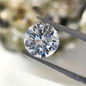 May include: A round, brilliant-cut clear gemstone held by a pair of metal tweezers. The stone features numerous triangular and kite-shaped facets that create a complex, symmetrical pattern of light reflection and refraction across its surface.