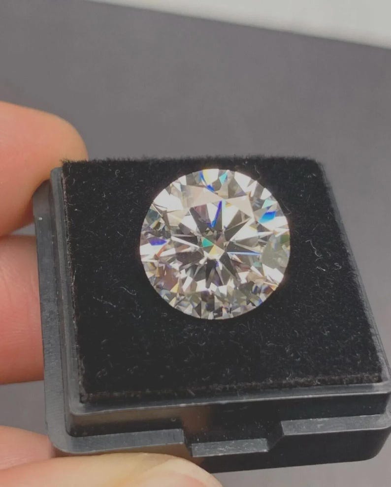 May include: A round, brilliant-cut, clear gemstone with multiple facets that reflect light, creating a sparkling effect with subtle rainbow-colored flashes. The gemstone is securely positioned within a square, black velvet-lined display box.