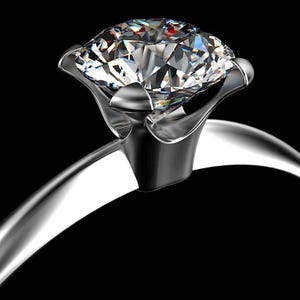 May include: A silver-toned ring featuring a large, round-cut, clear faceted gemstone set in a six-prong setting. The metal band is smooth and polished, reflecting light against a solid black background, highlighting the stone's brilliance.