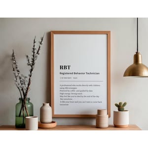 Puede incluir: Un marco de madera con el texto "RBT Registered Behavior Technician". El marco se exhibe en una estantería de madera con jarrones decorativos, un pequeño cactus y un jarrón de cristal con ramas.