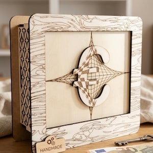 Puede incluir: Una hucha de madera hecha a mano con un diseño geométrico detallado. La caja presenta un acabado de madera clara con un marco cuadrado y un patrón decorativo. El diseño incluye una letra "E" estilizada y una etiqueta "HANDMADE".