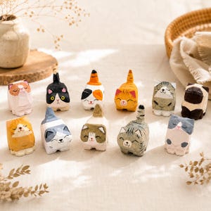Puede incluir: Una colección de doce pequeñas figuras de gatos talladas a mano en madera con diversos colores y patrones, incluyendo diseños atigrados, tricolores y sólidos. Cada gato presenta una expresión facial distinta, orejas erguidas y una pequeña cola.