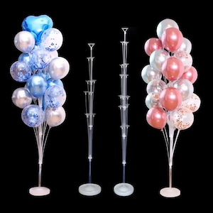 May include: A display of balloons in shades of blue, silver, and white, some with confetti, arranged on a stand. Two clear plastic balloon stands are also visible. The balloons are suitable for a party or celebration.