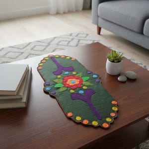 Puede incluir: Camino de mesa decorativo de fieltro verde con un diseño floral. El camino de mesa presenta una flor central con pétalos rojos, naranjas y azules, rodeada de hojas verdes. Acentos morados y puntos coloridos adornan los bordes.