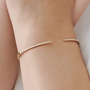May include: A gold-colored open cuff bracelet with a row of small, clear gemstones. The bracelet curves around the wrist, with the gemstones set along the top. The ends of the bracelet are open.