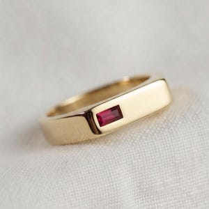 May include: A gold band ring featuring a single rectangular baguette-cut red gemstone set horizontally into the flat top surface. The metal has a smooth, polished finish, and the ring displays a clean, solid construction with a simple design.