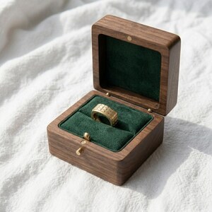 May include: A wooden ring box with a dark brown finish, open to reveal a gold ring on a green velvet interior. The ring has a textured design. The box is on a white surface.