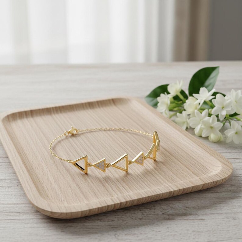 May include: A gold-colored bracelet with a series of triangle-shaped charms, each with a small, sparkling accent. The bracelet is displayed on a light wooden tray, with white flowers in the background.