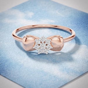 May include: Rose gold ring featuring a bow design with a cluster of small, sparkling diamonds at the center. The ring has a delicate band and is set against a light blue background.
