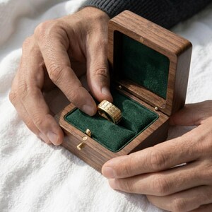 May include: A gold ring with a textured design rests in an open wooden ring box lined with green velvet. The ring box is held open by a person's hands, showcasing the ring's intricate details. The box is made of dark wood.
