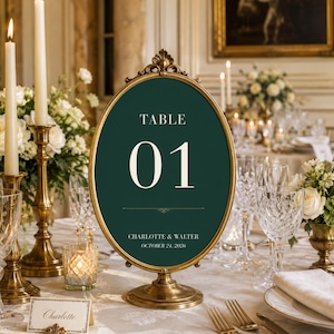 Op de afbeelding: Een ovaal tafelnummerbord in een goudkleurig metalen frame met een donkergroene achtergrond. Het bord toont de tekst TABLE 01, CHARLOTTE & WALTER en OCTOBER 24, 2026 in witte letters. Het bord is gemonteerd op een bijpassende gouden voet.