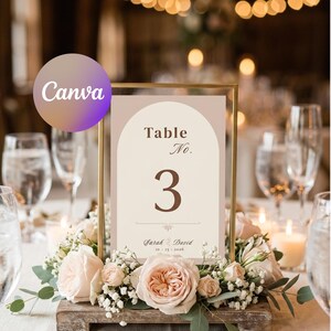 May include: A table number card with a gold frame, displaying "Table No. 3" in brown text. The card is surrounded by a floral arrangement of pink roses and greenery. The names "Sarah & David" and the date "10-25-2016" are also displayed.