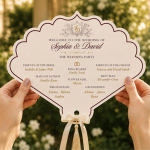 May include: A scalloped, fan-shaped wedding program card in cream with dark maroon borders. It features the text 'WELCOME TO THE WEDDING OF Sophia & David' and lists the wedding party members. A cream-colored ribbon and tassel are attached at the base.