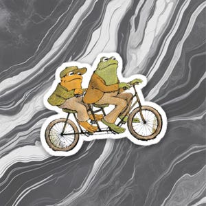 Op de afbeelding: Een sticker met twee antropomorfe kikkers op een tandemfiets. De ene kikker is oranje en draagt een groene jas en pet, de andere is groen en draagt een bruine jas. De fiets is bruin met witte banden, tegen een gemarmerde achtergrond.