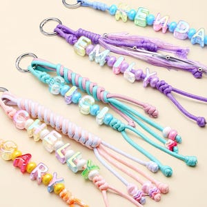May include: Five personalized keychains featuring braided cords in pastel colors, each attached to a silver metal ring. The keychains are decorated with iridescent letter beads spelling the names KENDALL, EMILY, CHELSEY, and MARY, interspersed with round beads.