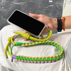May include: A smartphone in a clear case attached to a long, braided lanyard. The lanyard features a vibrant pattern of lime green and lavender paracord, finished with silver-toned metal end caps and adjustable knots for a secure, hands-free carry.