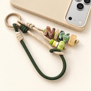 May include: A keyring featuring a dark green cord loop and a beige cord strand, connected by a beige plastic ring. The strand includes marbled brown, green, and beige letter beads spelling NINE, along with small green, yellow, and white accent beads.