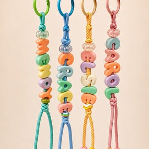 May include: Four colorful keychains hanging from a rope, each featuring a plastic ring, a knotted cord, and letter beads spelling the names Lennox, Grace, Elsie, and Carol. The keychains include various pastel-colored beads and small star charms.