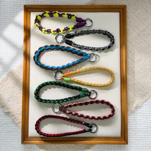 May include: A collection of colorful braided wrist straps with silver-tone key rings. The straps are in various colors, including yellow, purple, black, blue, green, pink, and red. The wrist straps are displayed in a wooden frame.