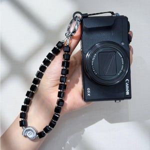 Könnte beinhalten: Eine schwarze Canon G5X Digitalkamera mit einer Handschlaufe aus facettierten schwarzen Würfelperlen und einem silberfarbenen Ammoniten-Anhänger. Das Objektiv zeigt den Text Canon Zoom Lens 5x IS sowie 8.8-44.0mm 1:1.8-2.8.