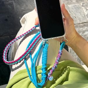 May include: A clear phone case with a blue, pink, and turquoise braided phone strap. The strap has a braided design with a turquoise base and pink and blue accents. The phone case has silver metal rings.