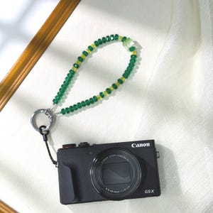Puede incluir: Una cámara Canon G5X negra con una correa de muñeca de cuentas verdes y amarillas. La correa está unida a un anillo de metal plateado. La lente de la cámara es visible y la correa está hecha de pequeñas cuentas de colores.
