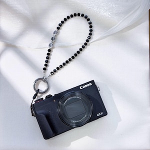 Pode incluir: Câmera Canon G5X preta com uma pulseira de contas pretas. A pulseira apresenta contas pretas e detalhes prateados. A lente da câmera está centralizada e o logotipo da Canon é visível.