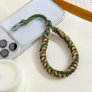 May include: A green and yellow braided phone wrist strap with a silver clasp. The strap is attached to a clear phone case. The wrist strap is made of a durable, woven material.