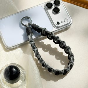 May include: A black and white braided phone wrist strap with a silver-tone metal ring. The strap is attached to a clear phone case. A black sphere sits in a clear glass container in the lower left corner.