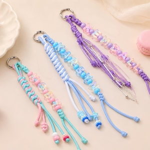 May include: Four handmade keychains featuring braided cord and colourful plastic beads. Each keychain includes a metal ring, knotted cord sections, and dangling strands with various bead shapes, including stars and letters in blue, purple, and pink.