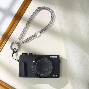 May include: Black Canon G5X camera with a beaded wrist strap. The strap features iridescent beads and a clear heart-shaped bead. The camera has a zoom lens and the Canon logo. The strap is attached to the camera with a black cord.