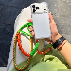 Puede incluir: Funda transparente para teléfono con un cordón trenzado verde y naranja. El cordón está sujeto a la funda del teléfono con anillos plateados. También se ve un reloj negro y una pulsera de cuentas.