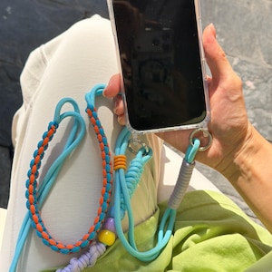 May include: A clear phone case with a blue lanyard. The lanyard has orange and blue braided sections, along with a gray, orange, and purple accent. The phone case is held by a person.