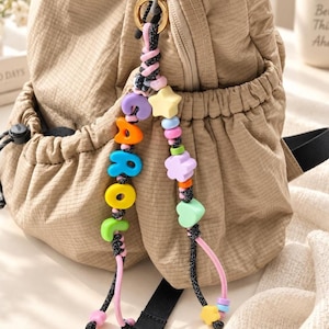 Può includere: Uno zaino beige con coulisse e un portachiavi decorativo di perline attaccato a un anello dorato. Il portachiavi è composto da due cordini neri con perline di plastica colorate, tra cui forme a stella, fiore, cuore e lettere che compongono CAROL.