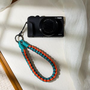 May include: A black Canon G5X camera with a teal and orange braided wrist strap. The strap is attached to the camera with a black cord and metal ring. The camera lens is visible.
