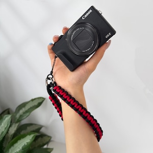 May include: A black Canon G5 X digital camera with a wrist strap. The strap is black and red, braided in a decorative pattern. The camera is held in a hand.
