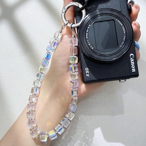 May include: A clear, iridescent crystal bead camera strap with silver-tone metal accents. The strap is attached to a black Canon camera. The beads are cube-shaped and reflect multiple colors.