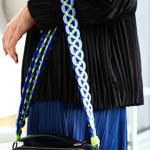 May include: A decorative bag strap featuring a braided design in vibrant shades of royal blue, light blue, and neon yellow. The strap is constructed from interconnected loops and attaches to a black rectangular bag with metallic hardware.