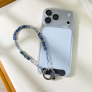 May include: A clear phone case with a blue beaded wrist strap. The strap features iridescent blue beads and a silver-coloured metal ring. The phone has a light blue screen and a black grip.