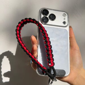 May include: A clear phone case with a black and red braided wrist strap. The strap is attached to the phone case with a silver-colored clasp. The phone case has three camera lenses.