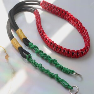 May include: A handmade camera strap featuring a red and white braided section, a black neck strap, and green braided wrist straps. The strap has yellow accents and silver metal rings for attachment.