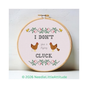 Può includere: Un telaio da ricamo a punto croce con la scritta "I DON'T give a flying cluck." Sono raffigurate due galline marroni, insieme a un bordo floreale e due pulcini gialli. Il telaio in legno è rotondo.