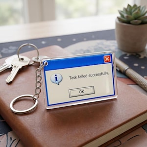 Op de afbeelding: Een heldere acryl sleutelhanger met een blauw en wit ontwerp dat lijkt op een computerfoutmelding. De tekst luidt "Task failed successfully." en bevat een "OK"-knop. De sleutelhanger is bevestigd aan een zilveren ketting en sleutelring, met sleutels zichtbaar.
