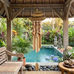 May include: A bamboo wind chime with a pagoda-style top hangs from a wooden structure. The chime features carved diamond patterns and cylindrical bamboo tubes. The background includes a pool, lush greenery, and a wooden bench, creating a serene outdoor setting.