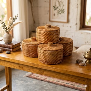 May include: Four round, woven rattan storage containers with lids sit on a wooden table. The containers are a warm, natural brown colour and have a textured surface. The lids have a small knob on top. The table is also made of wood.