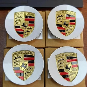 May include: Four white and gold Porsche wheel center caps. Each cap features the Porsche emblem with a black horse, gold accents, and red and black stripes. The caps are arranged on top of brown cardboard boxes.