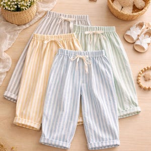 Puede incluir: Cuatro pares de pantalones de pijama a rayas en colores pastel: gris, amarillo, verde claro y azul. Cada par tiene una cintura con cordón. Un osito de peluche, sandalias y una cesta de flores también están en la imagen.